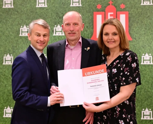 HFV-Präsident Christian Okun, Thomas Grage und HFV-Vizepräsidentin Kathrin Behn stehen gemeinsam vor einem Hintergrund mit HFV-Logos und zeigen eine Urkunde
